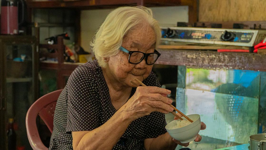 Angka Harapan Hidup Singapura Tembus 83 Tahun: 7 Rahasia & Tips 2025 1 Senior Asian woman eating rice with chopsticks in a cozy indoor setting.
