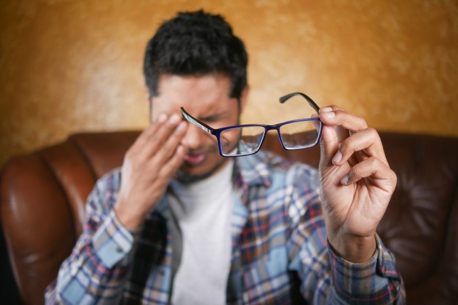 Awas! 9 Kebiasaan Tingkatkan Risiko Mata Minus & Cara Mencegahnya 1 A man in plaid shirt holding eyeglasses indoors. Focus on eyewear with neutral background.