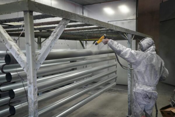 Worker in protective gear applying powder coating to metal pipes in a factory setting.
