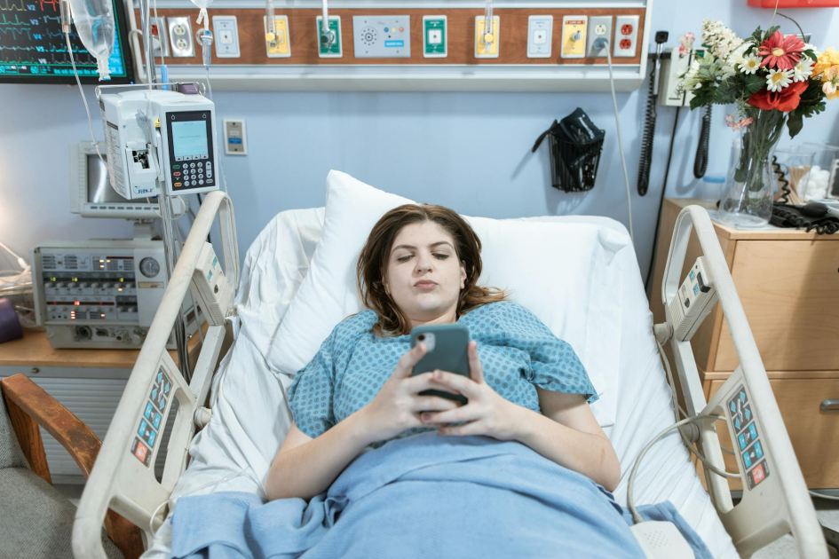 7 Fungsi Nurse Call dan Cara Kerjanya di Rumah Sakit 1 A female patient in a hospital bed using her phone, surrounded by medical equipment.