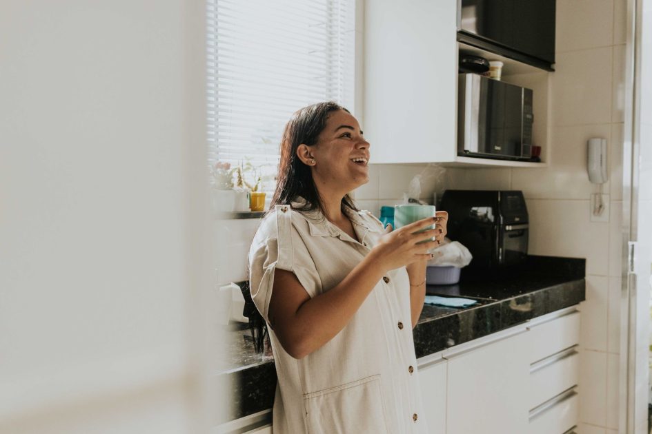Kata Dokter soal Tren Ngopi Gen Z: 7 Fakta & Solusi 2025 1 Relaxed woman standing in a kitchen, smiling and holding a cup of coffee, enjoying a sunny morning.