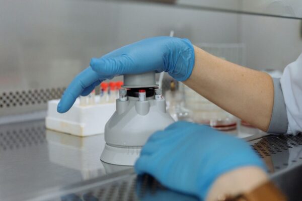 Close-up of a scientist's gloved hands operating centrifuge with laboratory equipment in the foreground.