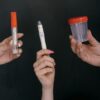 Three hands holding medical tools including a thermometer, lancet, and specimen container.