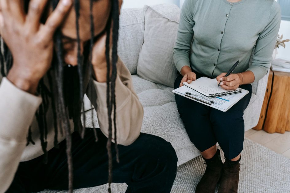 Crop concerned African American male sitting on sofa with female psychologist and holding head in hands in dismay during psychotherapy session
