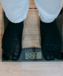 From above of crop unrecognizable plus size person in casual clothes standing on weighing scale on wooden floor