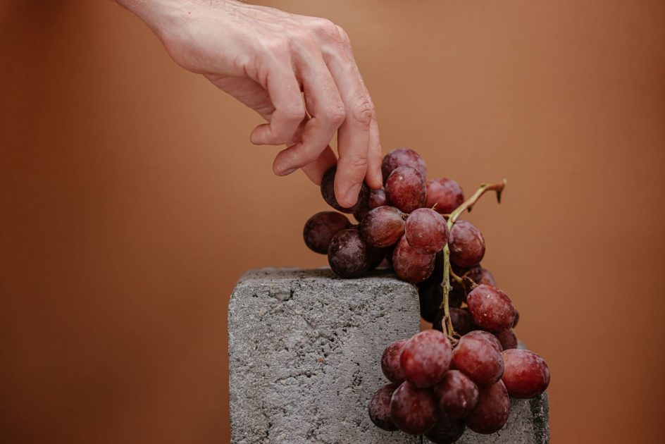 A close-up of a human hand picking juicy grapes from a stone block against a brown backdrop.
