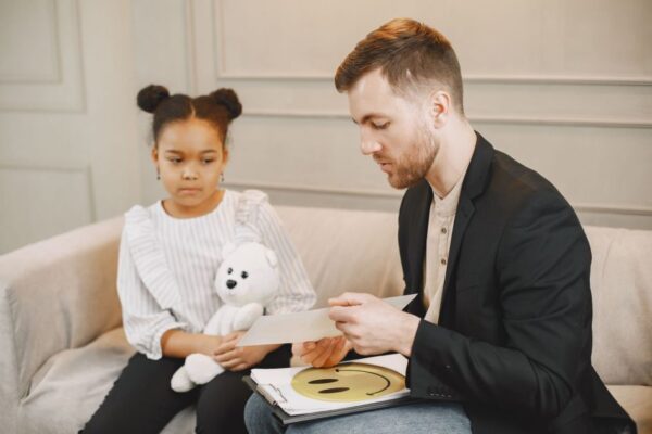 A child holding a plush toy listens to a man in a therapy session.