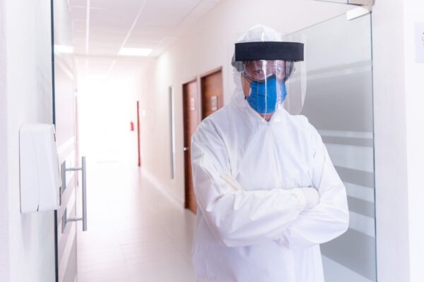 Medical professional in full PPE gear stands in a hospital corridor, wearing face mask and shield.