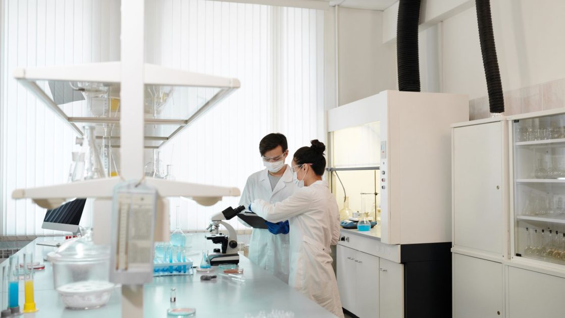 Particle Analyzer Farmasi: 6 Aplikasi Quality Control 1 Two scientists wearing protective gear collaborating in a laboratory setting with modern equipment.