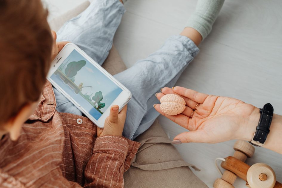 RI Hadapi Ancaman Silent Cognitive: 7 Strategi Deteksi Dini 1 Young child engrossed in a smartphone video while holding a brain model indoors.