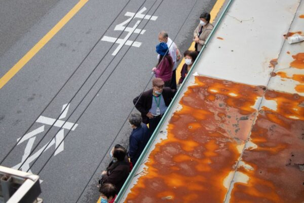 High angle view of people queuing at a bus stop with visible road markings and rusty roof.