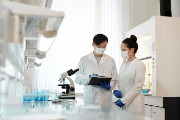 Two scientists in a laboratory wearing masks, discussing research with a microscope.