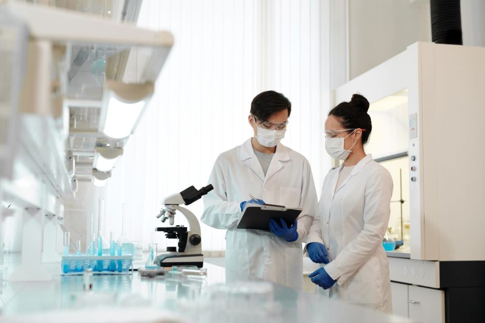 Two scientists in a laboratory wearing masks, discussing research with a microscope.