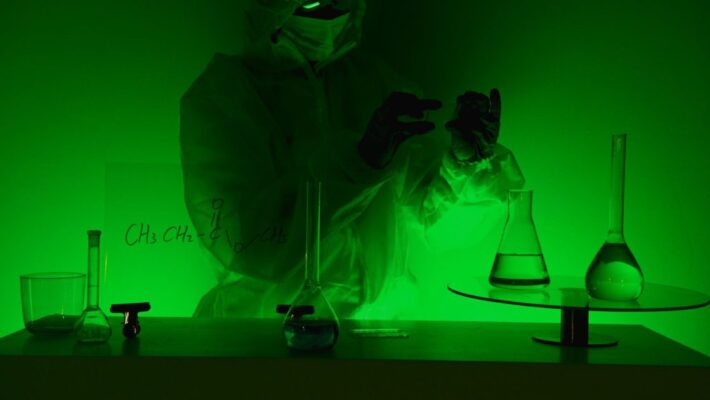 A scientist in protective gear works with glassware in a green-lit laboratory, evoking mystery and scientific exploration.