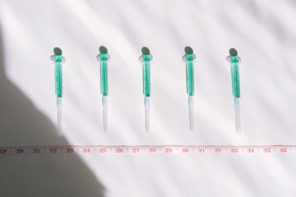 Top view of sterile syringes aligned with a centimeter ruler under bright light, casting shadows.