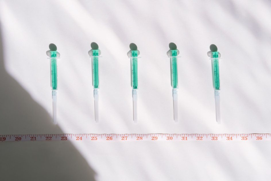 KPAK-KT Kepung Dinkes Kaltim: 7 Poin Transparansi Alkes 1 Top view of sterile syringes aligned with a centimeter ruler under bright light, casting shadows.