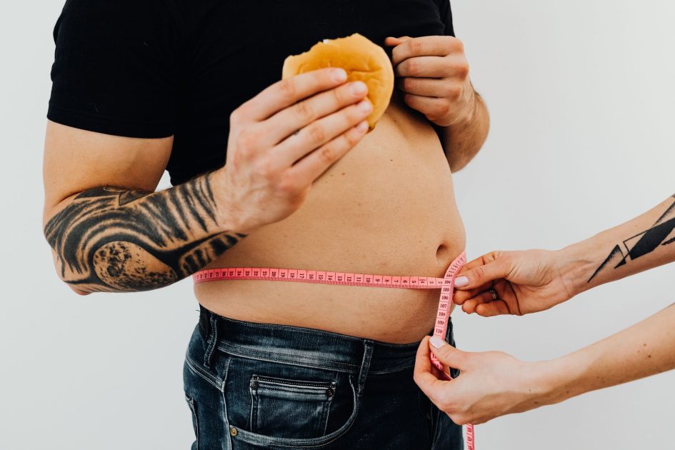 Kupas Tuntas Bahaya Obesitas & Produk Diet Ilegal 2025 1 A man with tattoos holding a burger as another person measures his waistline with a tape measure.