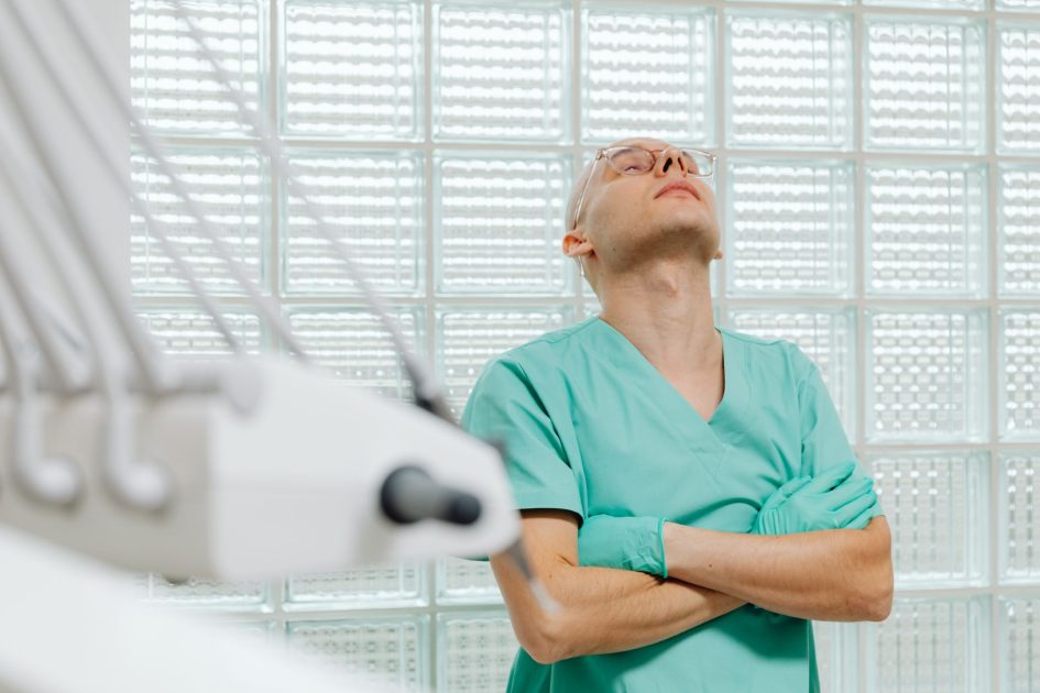 A healthcare worker in scrubs appears exhausted in a medical examination room.