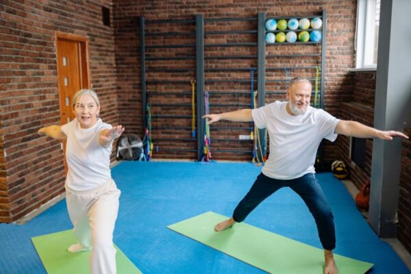 Elderly couple doing yoga poses on mats in a gym, promoting active lifestyle.