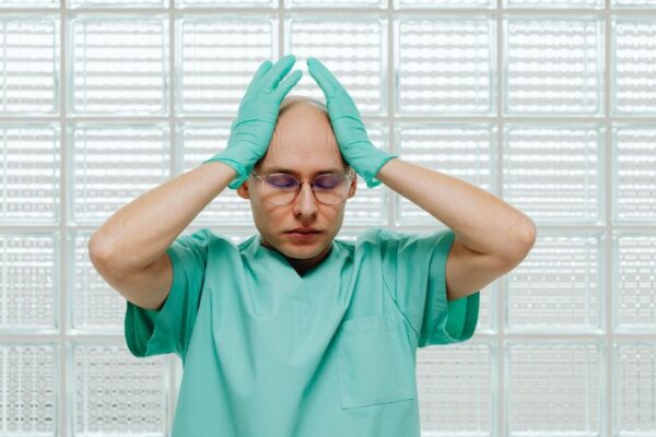 A healthcare worker in green scrubs looking stressed, hands on head with eyeglasses.
