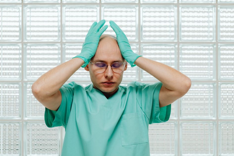 Rentetan Kematian Dokter Internsip Jadi Sorotan: 7 Faktor Penyebab & Solusi 1 A healthcare worker in green scrubs looking stressed, hands on head with eyeglasses.