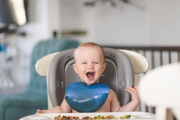 Happy baby laughing in a high chair during mealtime, wearing a blue bib.