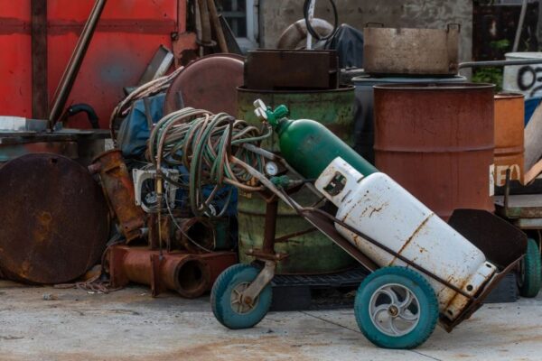 A cluttered industrial junkyard featuring gas cylinders, rusty drums, and tangled hoses.