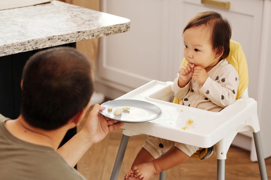 IDAI Desak Hentikan Keracunan MBG: 7 Langkah Pencegahan Penting 1 Adorable baby in a high chair eating while a parent offers food, indoors.