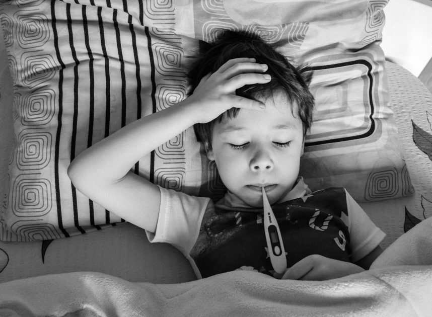 Influenza A Merebak: 7 Tanda Bahaya pada Anak & Panduan Penanganan 1 Black and white overhead view of child in casual clothing measuring body temperature with electronic thermometer while lying on bed under blanket