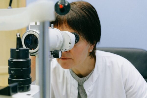 Anonymous female biologist in white coat observing object through big lab microscope while sitting on grey chair and leaning forward in light workplace