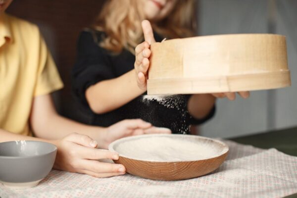 Crop cute girl and boy sifting flour through sieve in wooden plate for preparing dough while sitting at table covered with napkin on blurred background