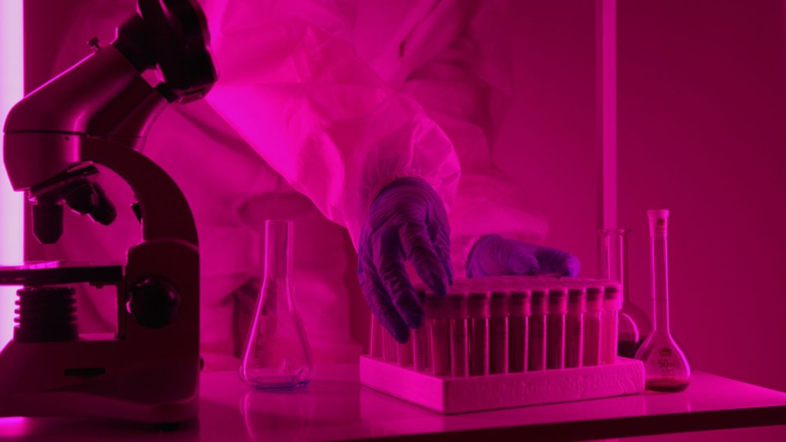 Bocah Meninggal Eksperimen Gen Editing: 7 Risiko & Pencegahan 1 Scientist in a lab coat handling test tubes under pink lighting, using a microscope.