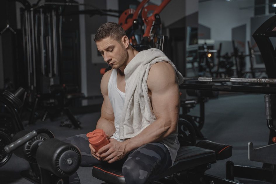 Butuh Recovery Sehabis Race? Ada 7 Metode Terbukti 2025 1 Muscular man sits in gym holding protein shaker after a workout, towel on shoulder.