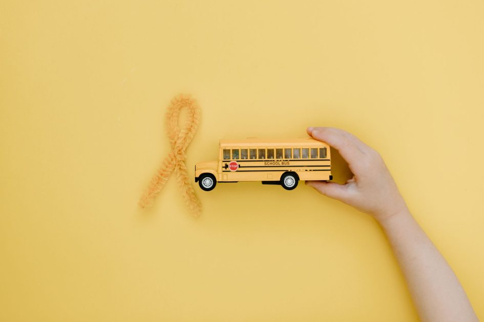 Hand holding toy school bus near yellow ribbon for cancer awareness on yellow background.
