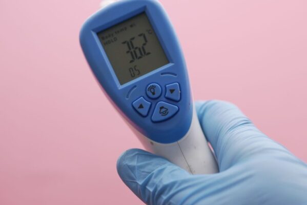 Close-up of an infrared thermometer displaying temperature, held by a gloved hand against a pink background.